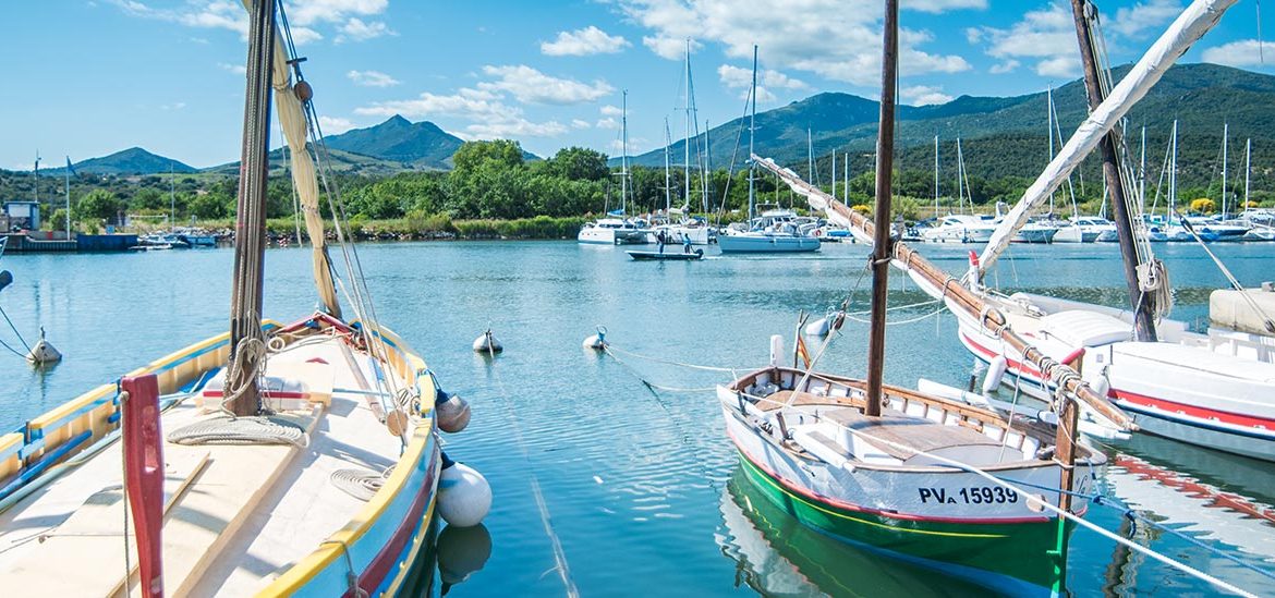 Des bateaux de plaisance amarrés dans un port de plaisance par une journée ensoleillée avec des montagnes en arrière-plan. Le ciel est bleu avec quelques nuages épars.