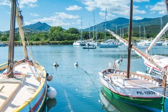 Des bateaux de plaisance amarrés dans un port de plaisance par une journée ensoleillée avec des montagnes en arrière-plan. Le ciel est bleu avec quelques nuages épars.