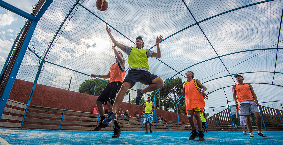 Joueurs de basketball en action sur un terrain multisport, portant des chasubles de couleurs vives sous un ciel partiellement nuageux.