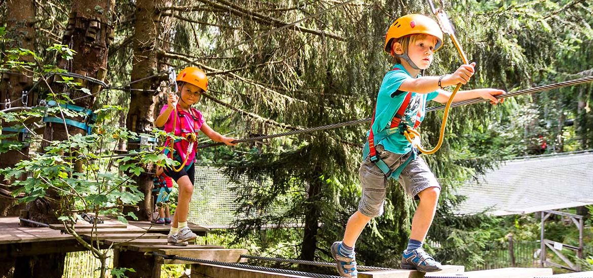 Deux enfants portant des casques et des harnais de sécurité traversant un parcours d'accrobranche en forêt.