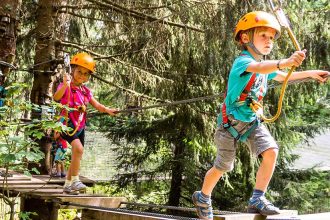 Deux enfants portant des casques et des harnais de sécurité traversant un parcours d'accrobranche en forêt.