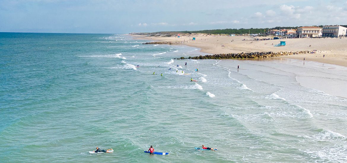 Surfeurs en train de prendre des vagues près du rivage sur une grande plage de sable avec quelques bâtiments en arrière-plan sous un ciel partiellement nuageux.