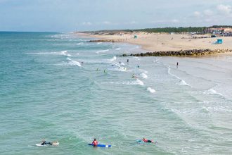 Surfeurs en train de prendre des vagues près du rivage sur une grande plage de sable avec quelques bâtiments en arrière-plan sous un ciel partiellement nuageux.