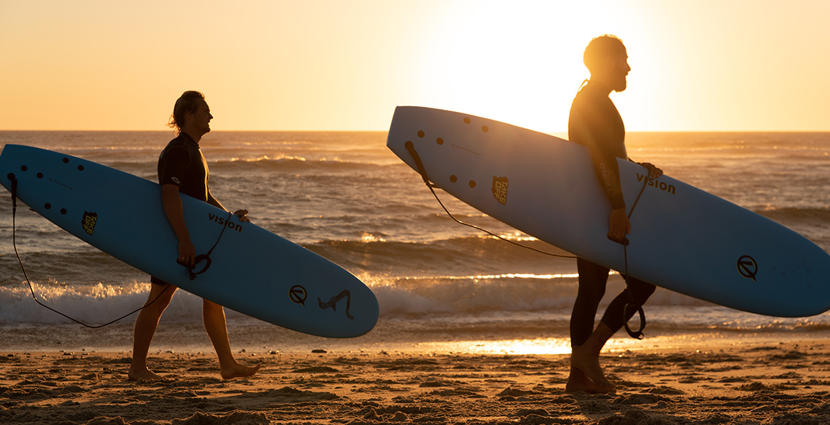 Deux surfeurs portant des planches bleues marchent sur la plage au coucher du soleil, avec l'océan en arrière-plan.