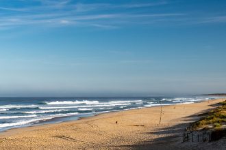 Longue plage de sable avec des vagues déferlantes sous un ciel bleu, entourée de dunes et de végétation côtière.
