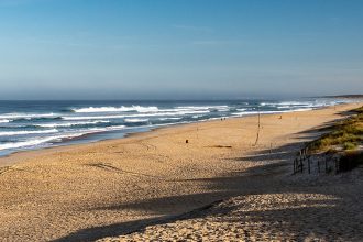 Longue plage de sable avec des vagues déferlantes sous un ciel bleu, entourée de dunes et de végétation côtière.