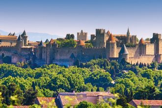 Vue panoramique de la cité médiévale de Carcassonne avec ses murs fortifiés, ses tours et ses bâtiments historiques entourés de verdure sous un ciel bleu clair.