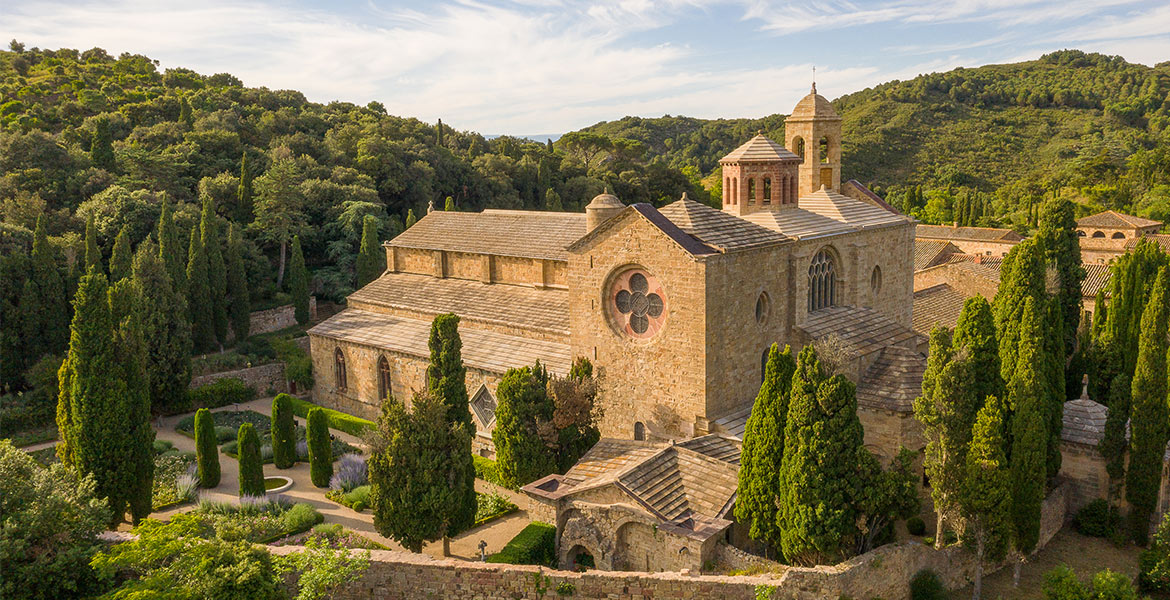 Vue aérienne de l'abbaye de Fontfroide dans l'Aude. C'est une abbaye médiévale en pierre, entourée de cyprès et de verdure, située dans une vallée boisée sous un ciel partiellement nuageux.