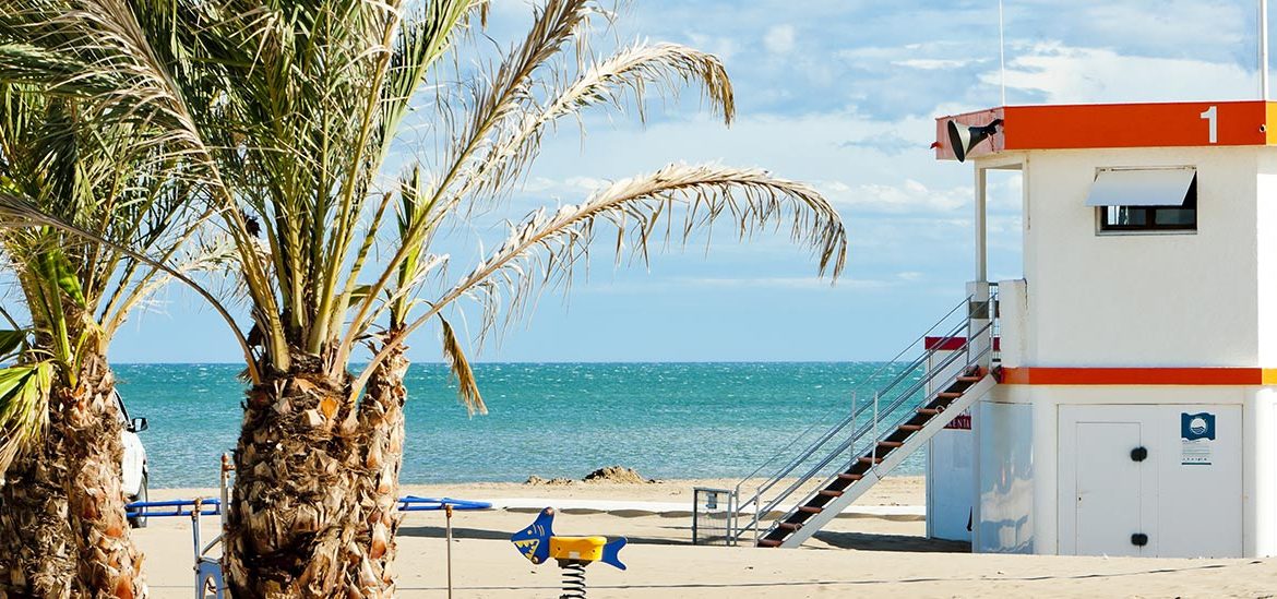 Poste de secours blanc et orange sur la plage de sable de Narbonne Plage, avec des palmiers au premier plan et la mer turquoise en arrière-plan sous un ciel partiellement nuageux.