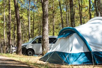 Un camping en forêt avec une grande tente blanche et bleue, des vans et des caravanes stationnés à proximité.