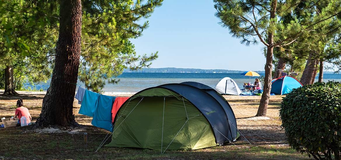 Un camping au bord de l'eau avec des tentes et du linge étendu entre les arbres. Une personne est assise sur le sol près de sa tente, profitant de la vue sur le lac ou la mer.