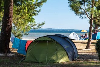 Un camping au bord de l'eau avec des tentes et du linge étendu entre les arbres. Une personne est assise sur le sol près de sa tente, profitant de la vue sur le lac ou la mer.