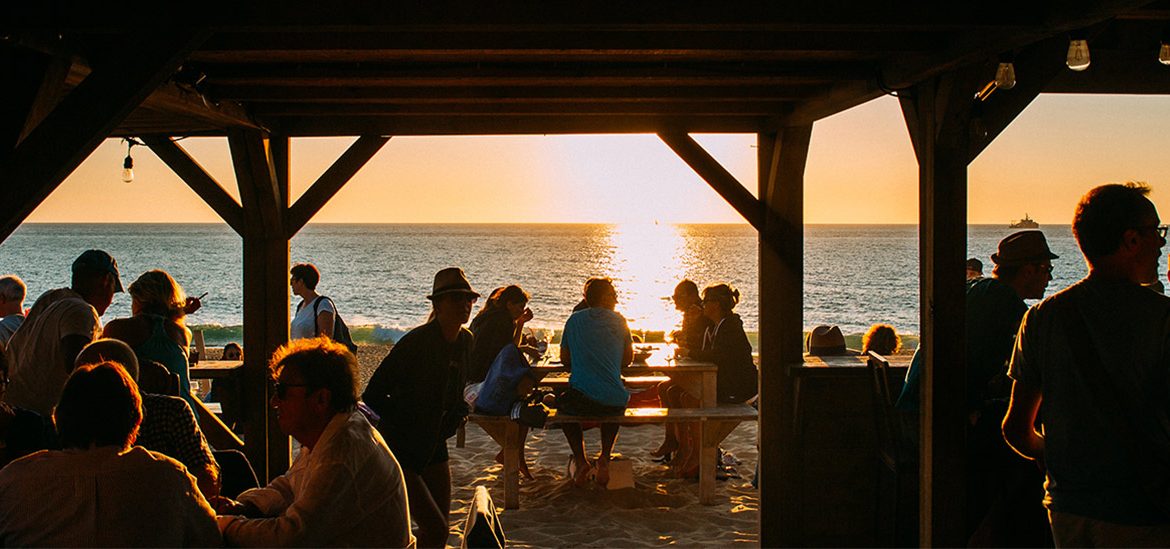 Des personnes assises sous une structure en bois sur la plage, profitant d'un coucher de soleil avec vue sur la mer.