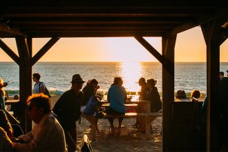 Des personnes assises sous une structure en bois sur la plage, profitant d'un coucher de soleil avec vue sur la mer.