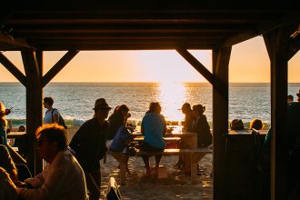 Des personnes assises sous une structure en bois sur la plage, profitant d'un coucher de soleil avec vue sur la mer.