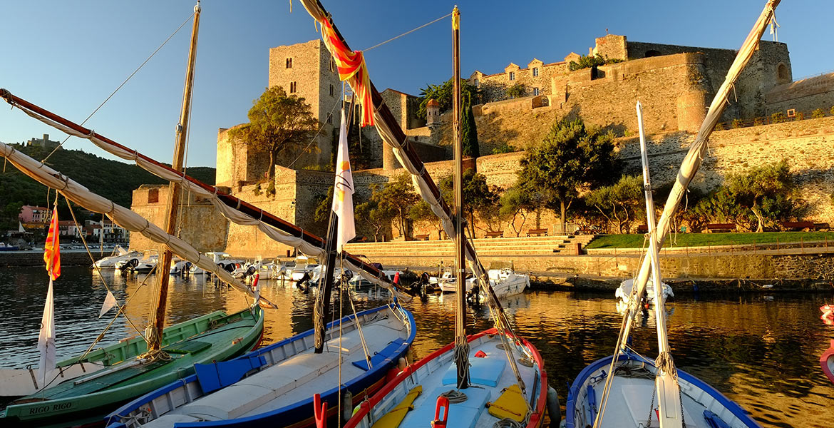 Des bateaux colorés sont amarrés dans un port devant une ancienne forteresse en pierre, illuminée par la lumière dorée du coucher de soleil. Les eaux calmes reflètent les voiles et les murs historiques.