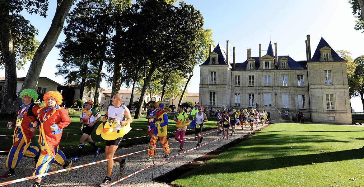 Des coureurs déguisés en clowns et autres costumes colorés participent à une course devant un château majestueux, entouré de jardins et d'arbres.