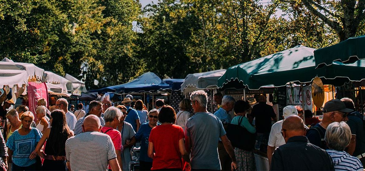 Un marché en plein air animé avec des stands couverts de toiles colorées, des gens faisant leurs achats et des arbres en arrière-plan.