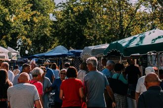 Un marché en plein air animé avec des stands couverts de toiles colorées, des gens faisant leurs achats et des arbres en arrière-plan.