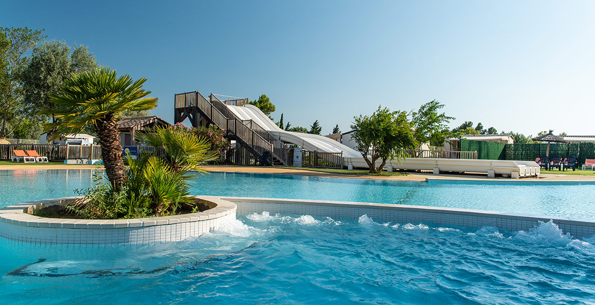 Piscine extérieure du Camping Campéole La Côte des Roses à Narbonne Plage dans l'Aude avec bains à remous, palmier sur îlot central, toboggan aquatique en arrière-plan avec ciel bleu dégagé.