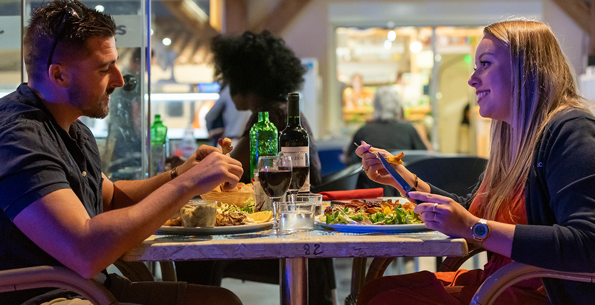 Un homme et une femme souriants en train de dîner dans un restaurant au Camping Campéole La Côte des Roses à Narbonne Plage dans l'Aude, avec des assiettes de nourriture et une bouteille de vin sur la table.