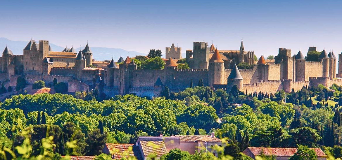 Vue panoramique de la cité médiévale de Carcassonne avec ses imposants remparts et tours, entourée de verdure. Le ciel bleu clair contraste avec la végétation luxuriante au premier plan.