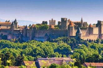 Vue panoramique de la cité médiévale de Carcassonne avec ses imposants remparts et tours, entourée de verdure. Le ciel bleu clair contraste avec la végétation luxuriante au premier plan.