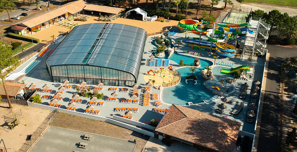 Vue aérienne d'un complexe aquatique avec piscines extérieures, toboggans et espace détente avec des transats sous des parasols.