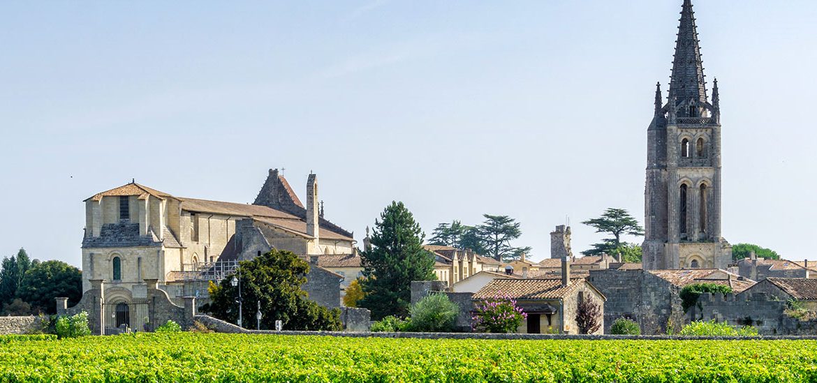 Vue sur un vignoble verdoyant avec le village de Saint-Émilion en Gironde et une église à clocher pointu en arrière-plan sous un ciel bleu légèrement nuageux.