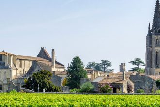 Vue sur un vignoble verdoyant avec le village de Saint-Émilion en Gironde et une église à clocher pointu en arrière-plan sous un ciel bleu légèrement nuageux.