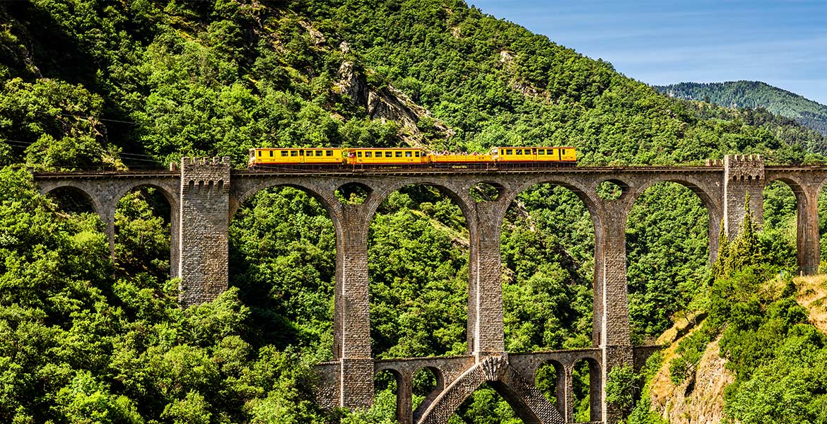 Le train jaune des Pyrénées-Orientales traversant un grand viaduc en pierre au milieu d'un paysage boisé.