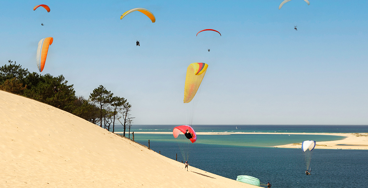 Parapentistes colorés volant au-dessus de la Dune du Pilat près de la côte, avec vue sur l'océan et une plage au loin.