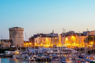 Panorama du crépuscule sur le port de La Rochelle avec les Tours de la Chaîne et Saint-Nicolas, des bateaux amarrés et l'illumination des façades et des monuments historiques en arrière-plan.