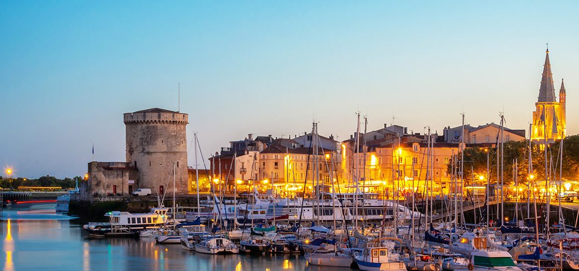 Panorama du crépuscule sur le port de La Rochelle avec les Tours de la Chaîne et Saint-Nicolas, des bateaux amarrés et l'illumination des façades et des monuments historiques en arrière-plan.