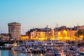 Panorama du crépuscule sur le port de La Rochelle avec les Tours de la Chaîne et Saint-Nicolas, des bateaux amarrés et l'illumination des façades et des monuments historiques en arrière-plan.