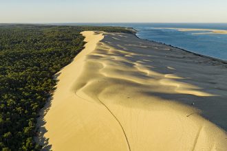 Vue aérienne de la Dune du Pilat séparant une forêt dense et une plage, avec l'océan à l'horizon sous un ciel clair.