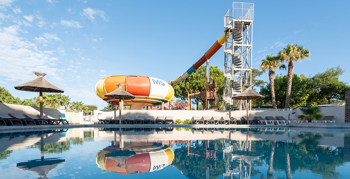 Piscine avec des chaises longues et parasols en paille, avec un toboggan aquatique coloré et une tour de glissade en arrière-plan. Le ciel est dégagé, reflétant sur l'eau calme de la piscine.