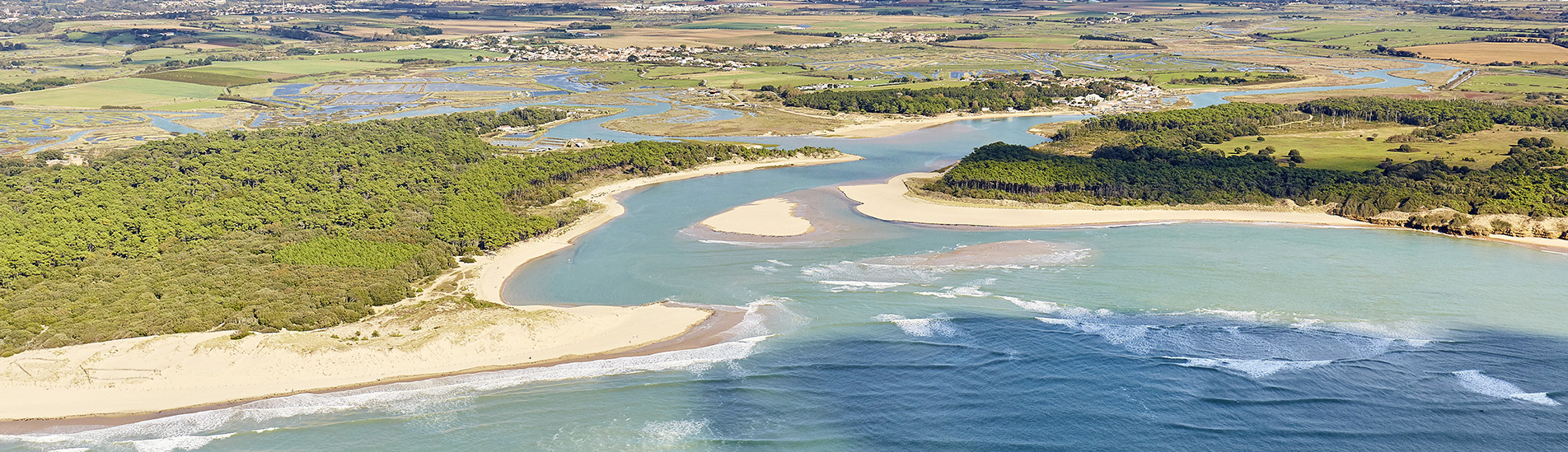 Vue aérienne d'une côte avec des plages de sable, des eaux bleu turquoise et des zones humides, entourée de terres agricoles et de quelques bâtiments.