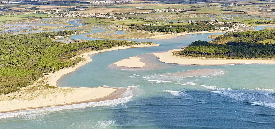 Vue aérienne d'une côte avec des plages de sable, des eaux bleu turquoise et des zones humides, entourée de terres agricoles et de quelques bâtiments.