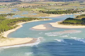 Vue aérienne d'une côte avec des plages de sable, des eaux bleu turquoise et des zones humides, entourée de terres agricoles et de quelques bâtiments.