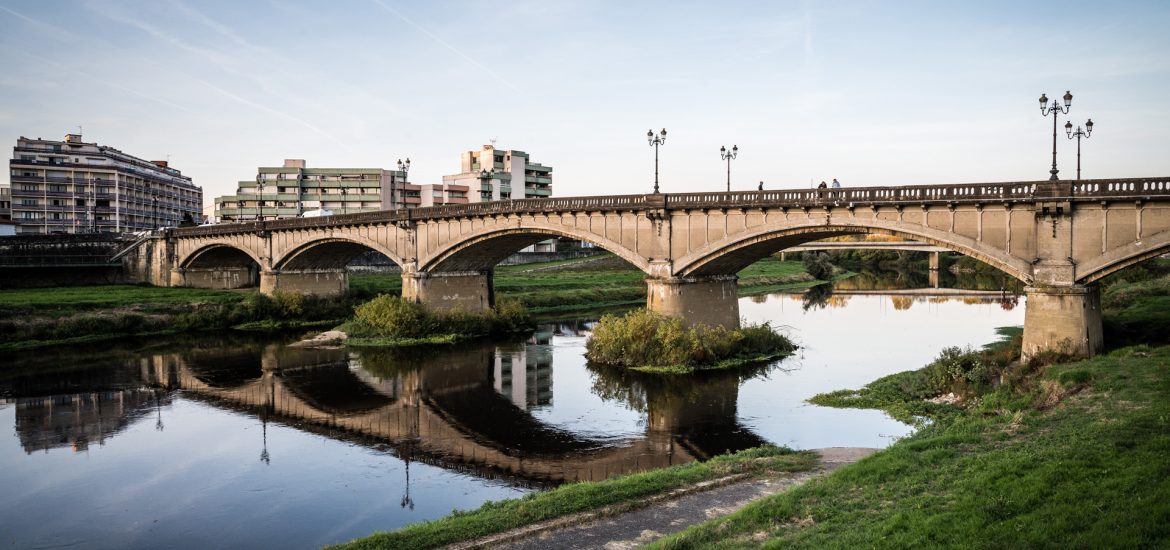 Pont en pierre de Dax avec arches se reflétant dans une rivière, entouré de bâtiments résidentiels modernes en arrière-plan, sous un ciel clair.
