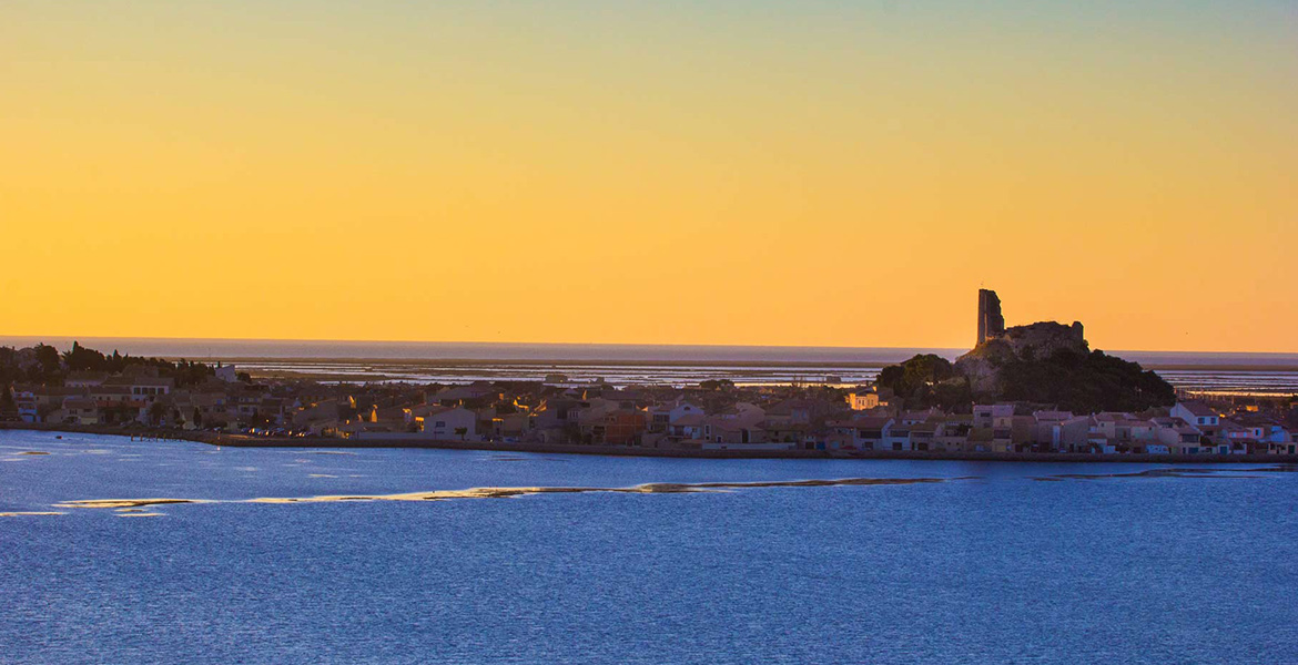 Coucher de soleil sur un village côtier avec une ancienne tour en ruine sur une colline surplombant la mer.