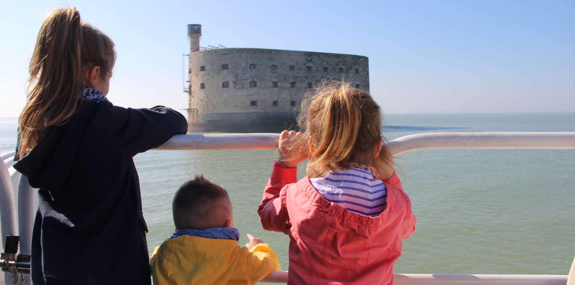 Trois enfants de dos regardant le Fort Boyard depuis le pont d'un bateau en mer par une journée ensoleillée.