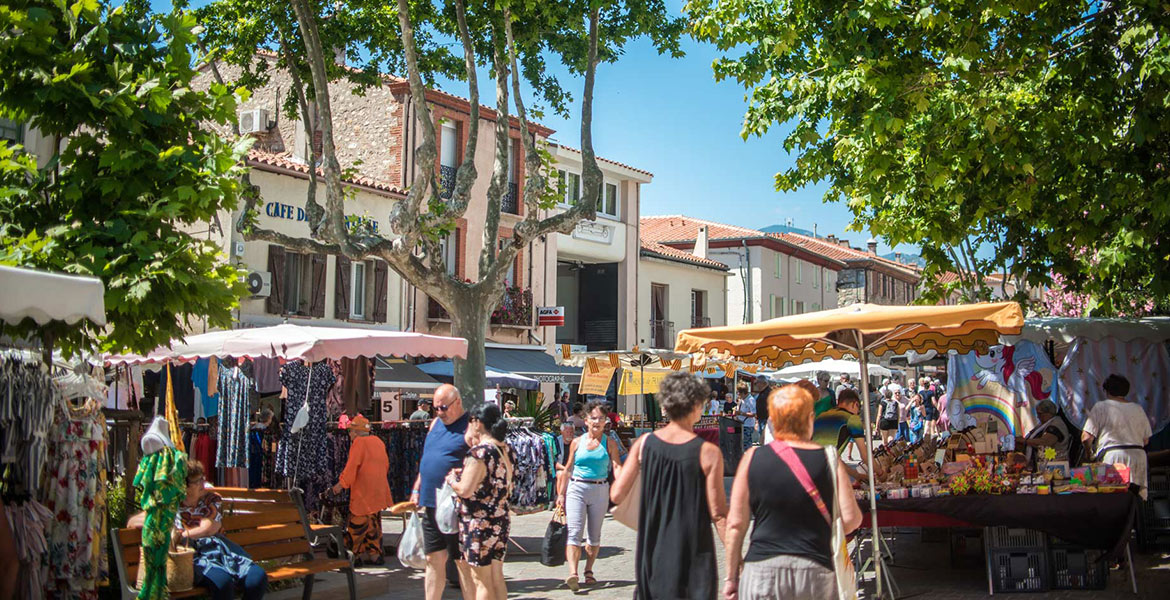 Marché en plein air de la ville d'Argeles sur mer, avec des étals colorés sous des auvents, des vêtements suspendus, et des gens déambulant parmi les stands. En arrière-plan, des bâtiments en pierre et des arbres fournissent de l'ombre.