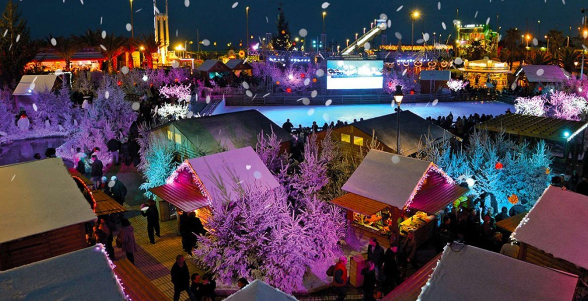 Marché de Noël de Barcarès en soirée avec des chalets illuminés, des arbres décorés de lumières colorées, et une patinoire au centre. De la neige artificielle tombe, ajoutant à l'ambiance festive. Des gens se promènent et visitent les stands.