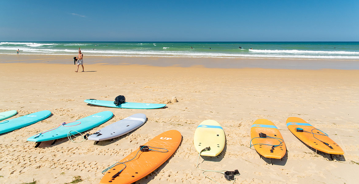 Plage avec plusieurs planches de surf colorées alignées sur le sable, des surfeurs dans l'eau et un homme marchant sur le rivage sous un ciel bleu clair.