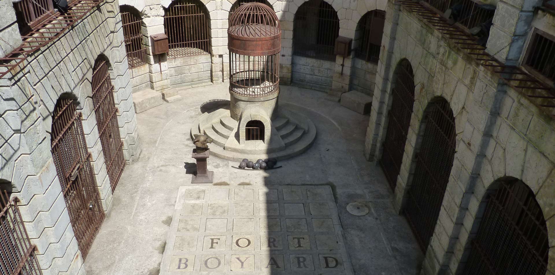 Vue intérieure de la cour centrale de Fort Boyard, avec une cage en métal au centre et les mots "Fort Boyard" gravés sur le sol en dalles de pierre.