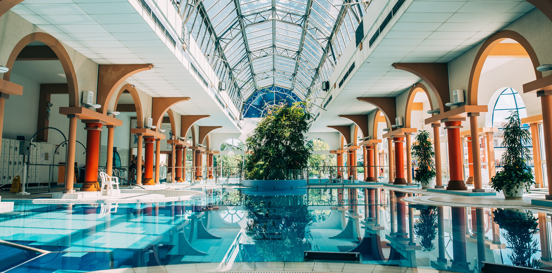 Les thermes sont une activité à faire à Dax. Une piscine intérieure avec des arches et un toit en verre, entourée de plantes et de colonnes, éclairée par la lumière naturelle.