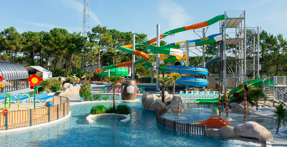 Aire de jeux aquatiques avec des toboggans colorés et des bassins peu profonds, entourée de chaises longues et de verdure. Des structures de jeux pour enfants sont installées près de l'eau.