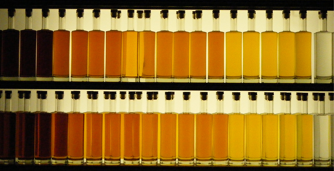 Deux rangées de bouteilles remplies de différentes nuances de cognac sont alignées sur une étagère. Les couleurs varient du brun foncé à l'ambre clair, montrant une gamme de vieillissement.